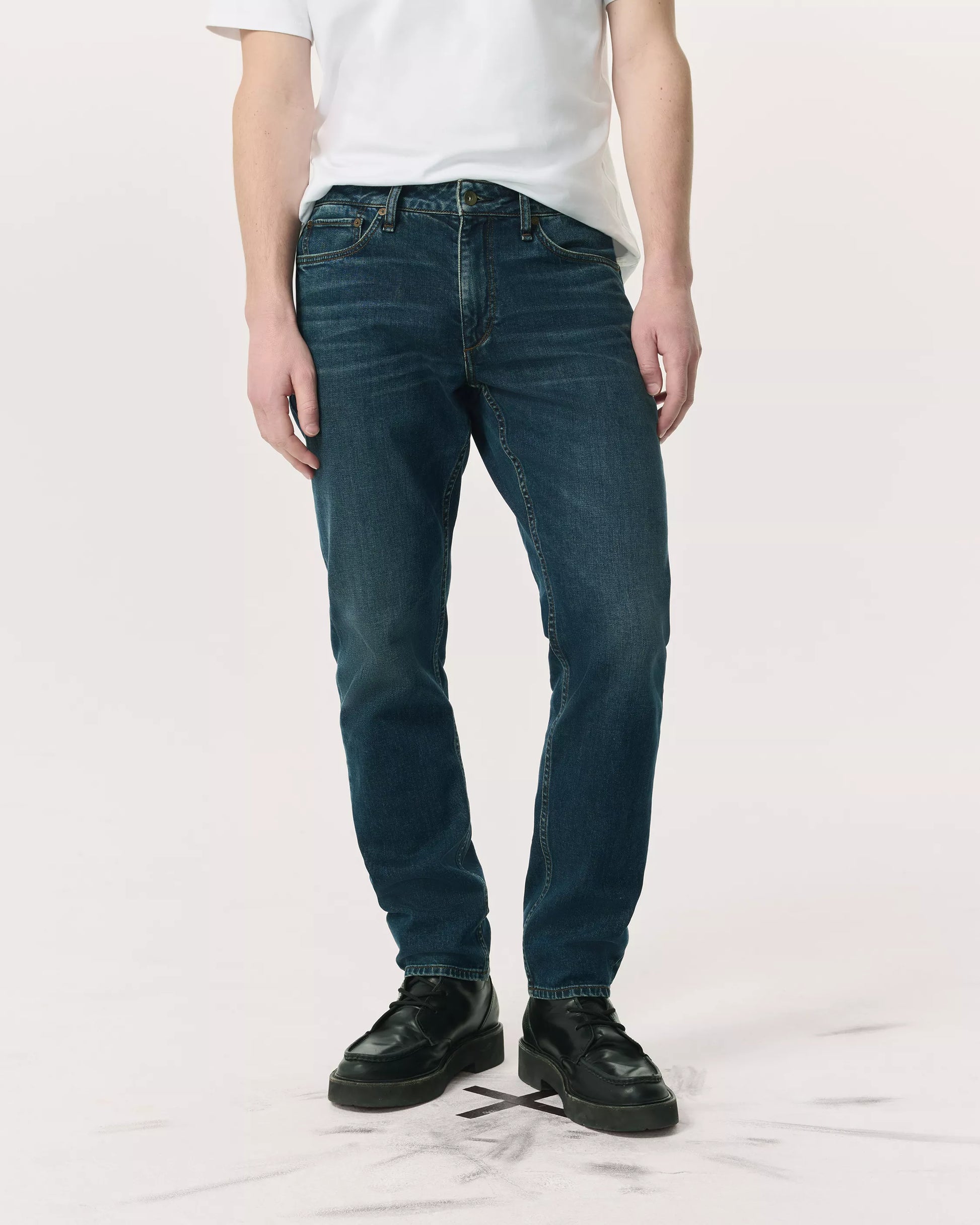 A person wearing a white T-shirt, Rag & Bone Fit 2 Authentic Stretch Denim jeans in dark indigo, and black shoes stands against a plain light background.