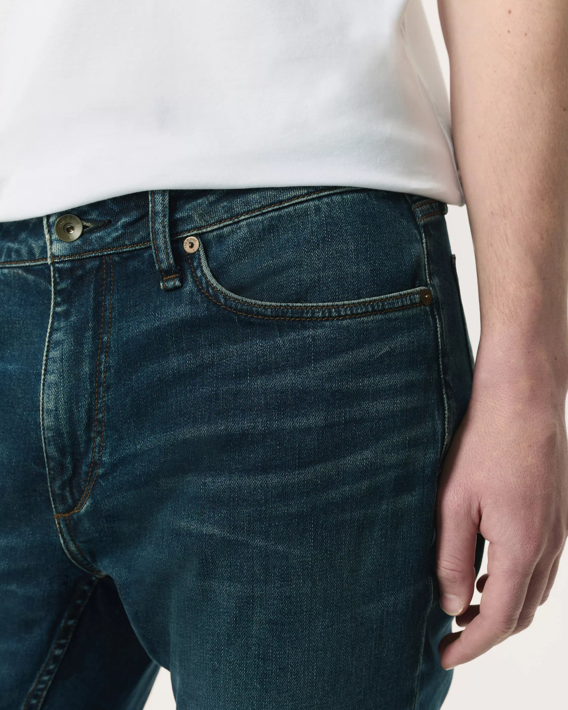 A person wearing Rag & Bone Fit 2 Authentic Stretch Denim jeans in a dark indigo wash and a white shirt, with their right hand resting by their side.