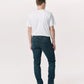 A man stands facing away, wearing a plain white t-shirt, black shoes, and Rag & Bone Fit 2 Authentic Stretch Denim jeans in a dark indigo wash, set against a light neutral background.