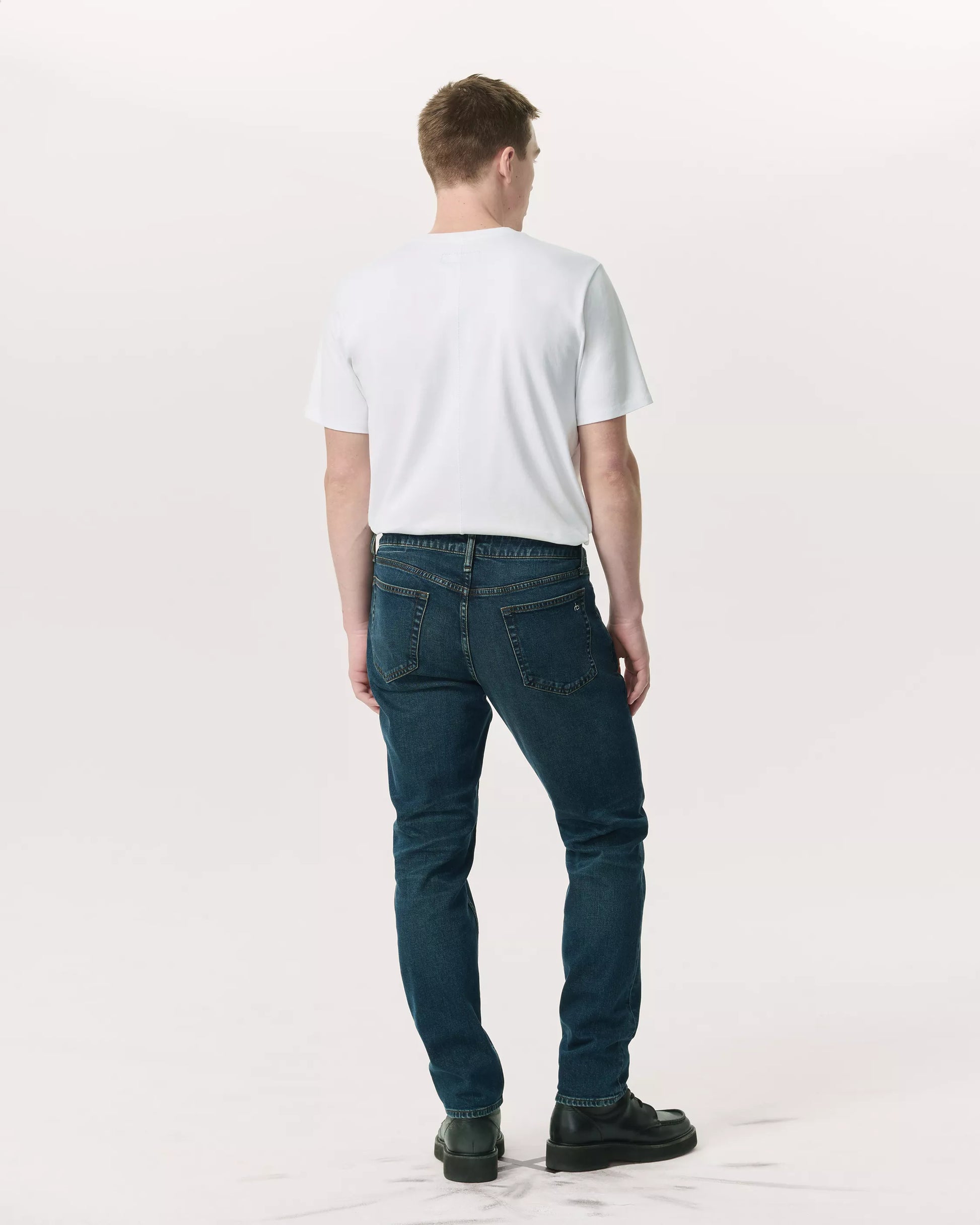 A man stands facing away, wearing a plain white t-shirt, black shoes, and Rag & Bone Fit 2 Authentic Stretch Denim jeans in a dark indigo wash, set against a light neutral background.