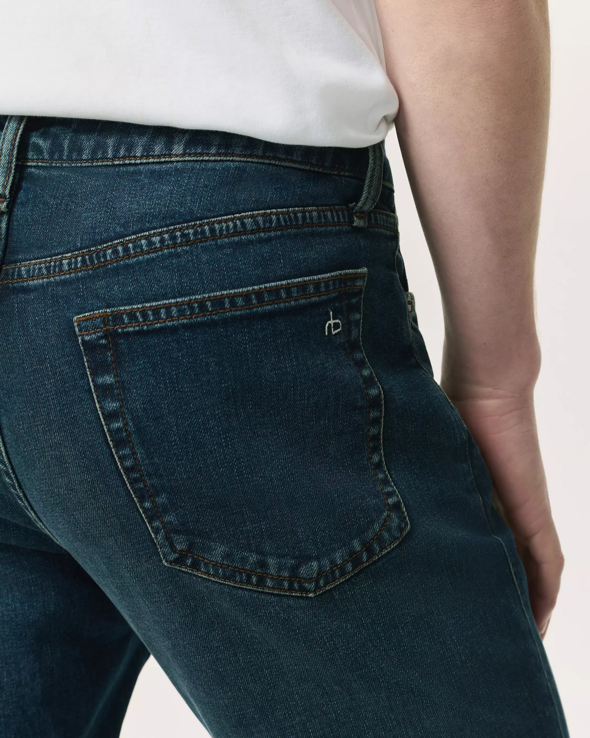 A person wears Rag & Bone Fit 2 Authentic Stretch Denim jeans in dark indigo, paired with a white shirt, shown from the waist down with emphasis on the back pocket.