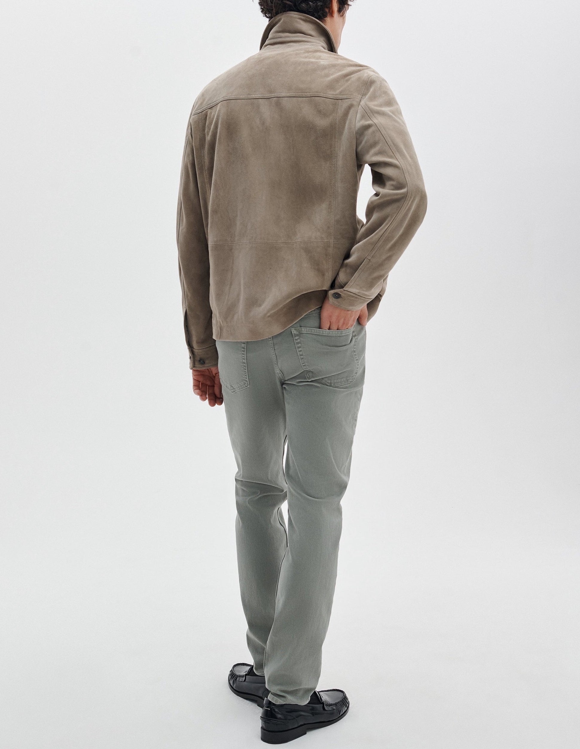A man faces away against a white background, wearing a taupe suede jacket, Rag & Bone Fit 2 Aero Stretch Denim light gray pants, and black shoes.