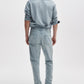 A person stands facing away, dressed in a light blue button-up shirt, Rag & Bone Fit 3 Authentic Stretch athletic fit jeans with a tapered leg in light blue, and black shoes against a plain white background.