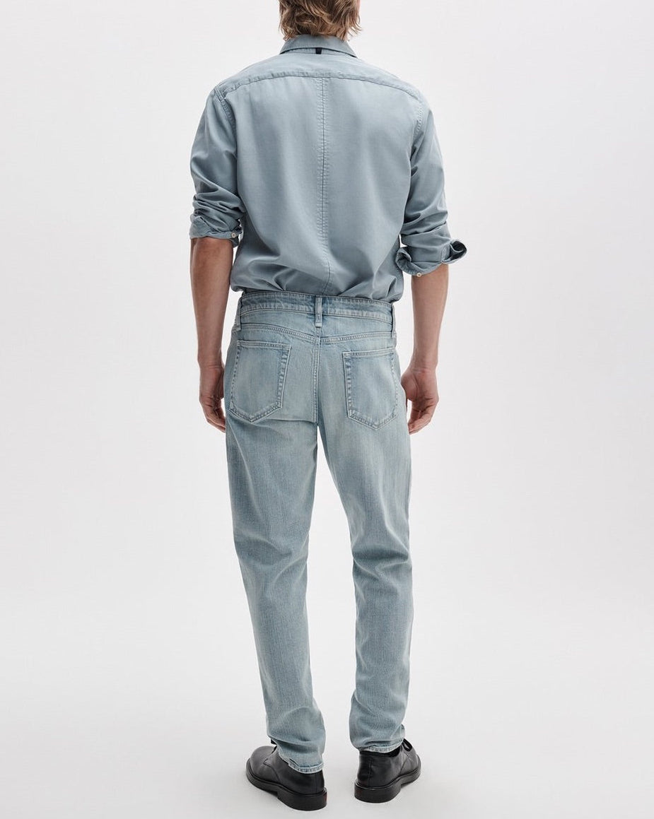 A person stands facing away, dressed in a light blue button-up shirt, Rag & Bone Fit 3 Authentic Stretch athletic fit jeans with a tapered leg in light blue, and black shoes against a plain white background.