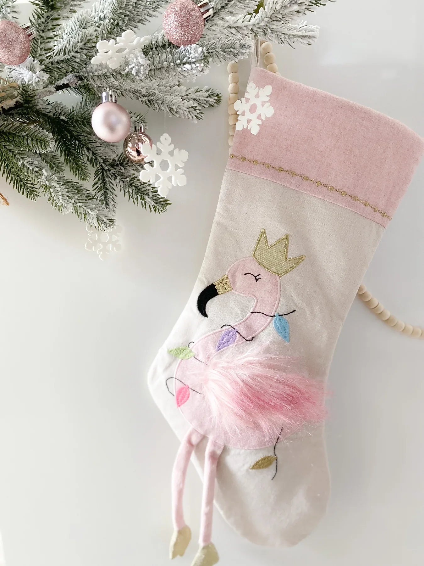 Introducing the Mon Ami Festive Flamingo Stocking: This delightful pink stocking, adorned with a regal flamingo embroidered with a gold crown, is beautifully decorated with colorful Christmas lights and intricate designs. It makes for a charming festive decoration when hung among evergreen branches, delicate snowflakes, and baubles.