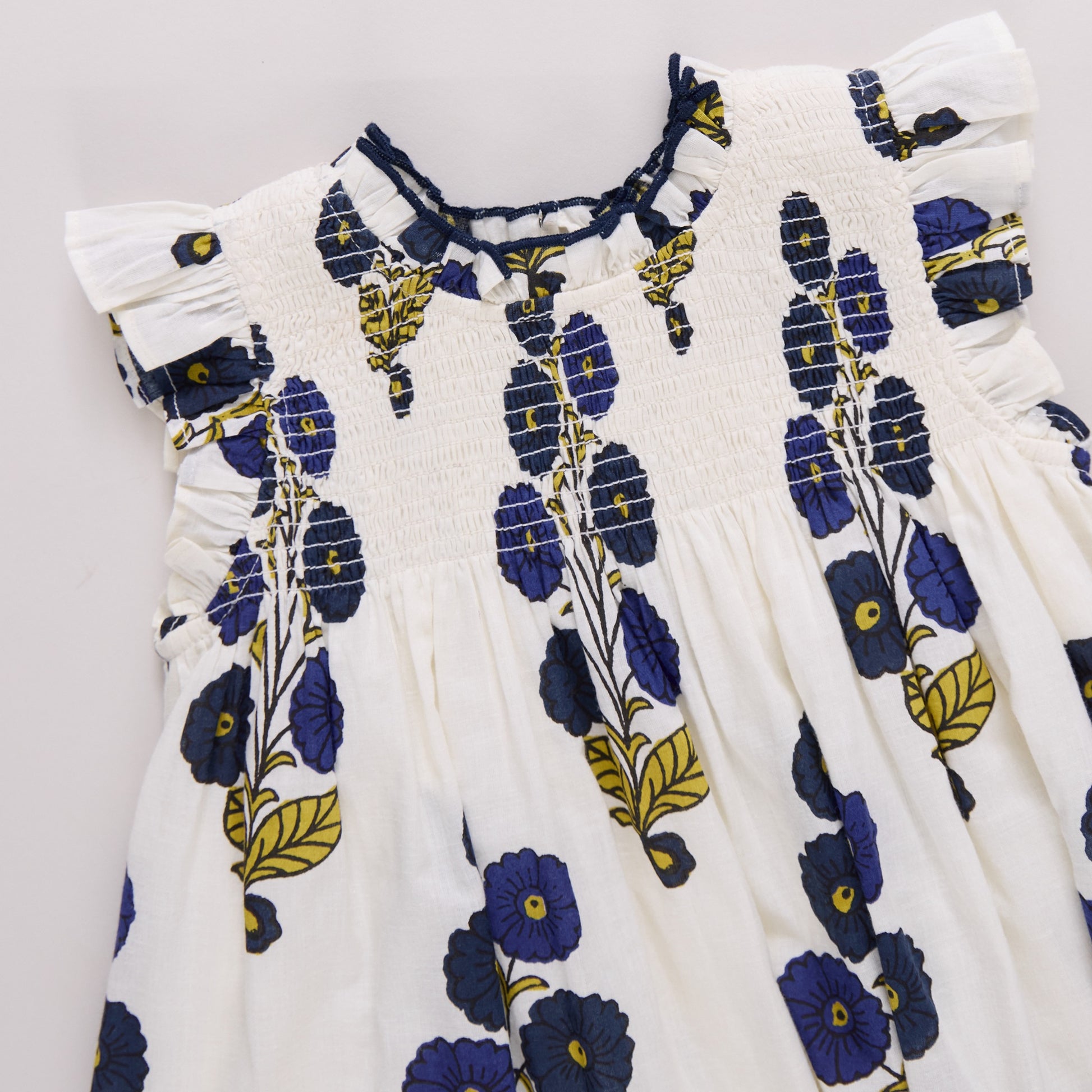The Pink Chicken Girls' Stevie Dress features a white base with blue and yellow floral print, short ruffled sleeves, and shirred chest detailing for a charming look.