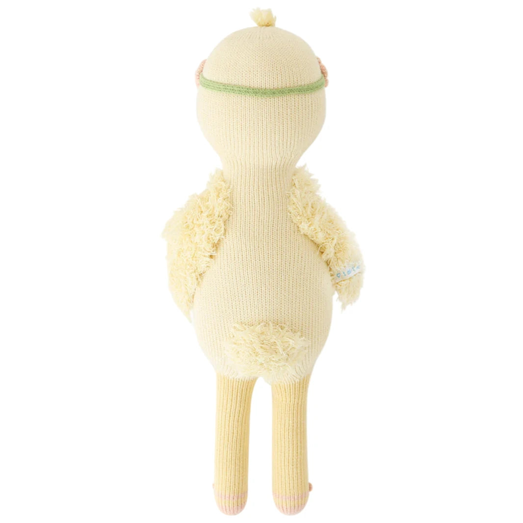 Flora the Duckling, Mauve - Regular 20" by Cuddle + Kind is a knitted plush toy shown from the back, with textured wings and a tuft on its head, standing as if dancing near a sparkling river amid imaginary wildflowers.