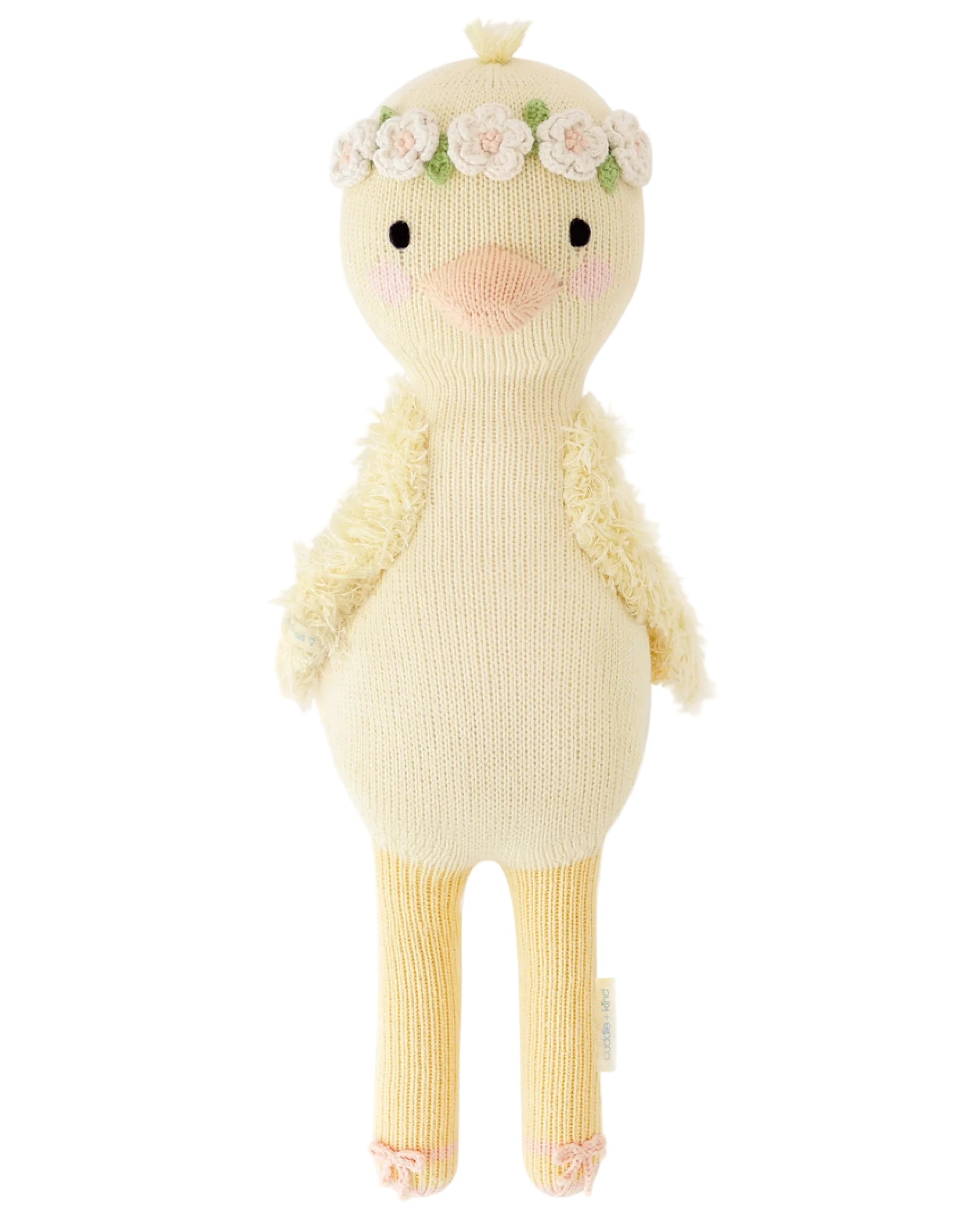 Flora the Duckling Ivory by Cuddle + Kind is a 20" stuffed toy featuring long legs, textured wings, and a floral headband, standing upright as if dancing among wildflowers against a white background.