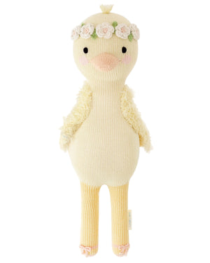 Flora the Duckling Ivory by Cuddle + Kind is a 20" stuffed toy featuring long legs, textured wings, and a floral headband, standing upright as if dancing among wildflowers against a white background.