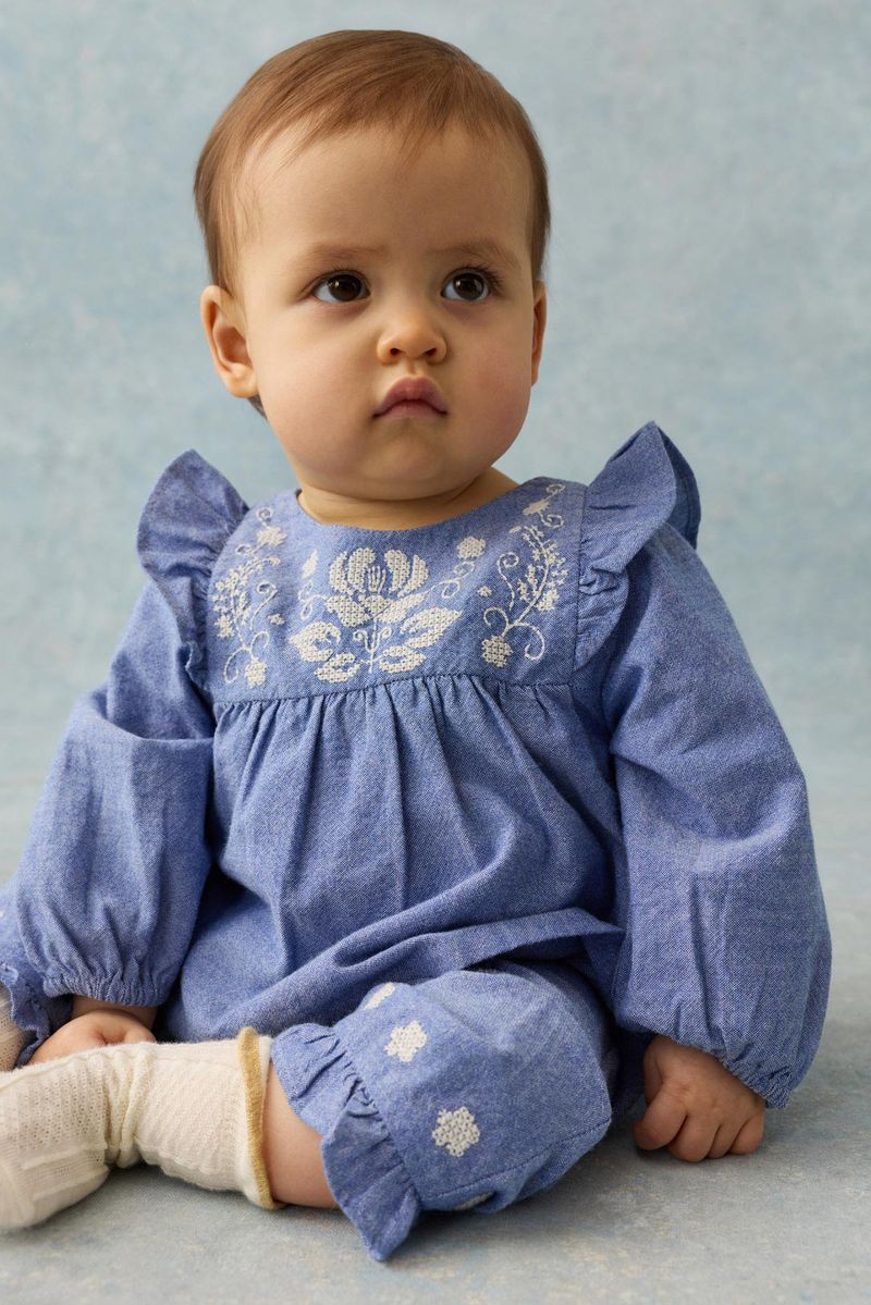A baby wearing the Louise Misha Baby Girls’ Mikani Set—an embroidered blue dress with white floral details—sits on a blue background, looking slightly to the side with a neutral expression.
