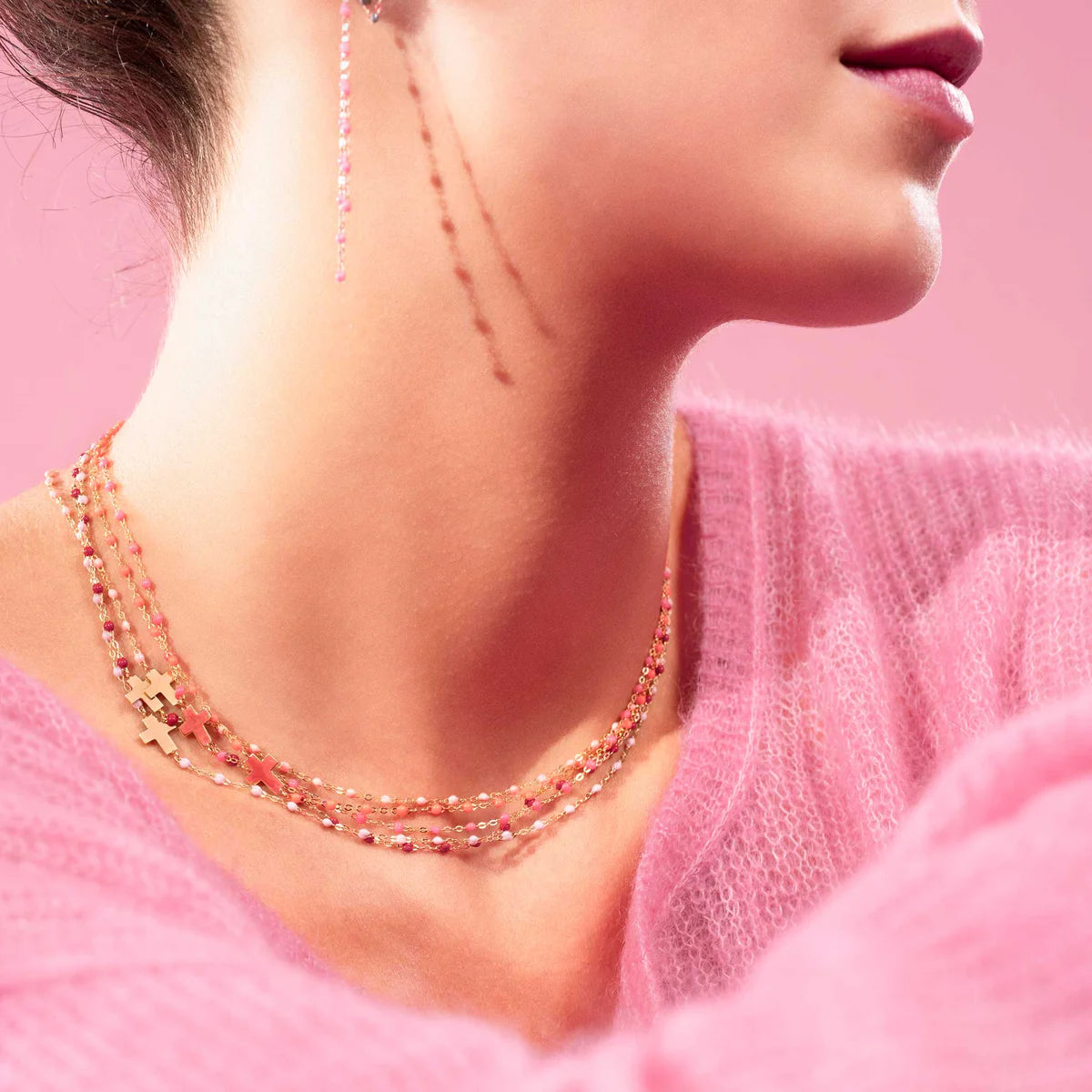 A person wearing a pink fuzzy sweater, Gigi Clozeau’s Side Cross Necklace 16.5" with resin pearls and 18k gold, plus matching dangle earrings, poses against a pink background.