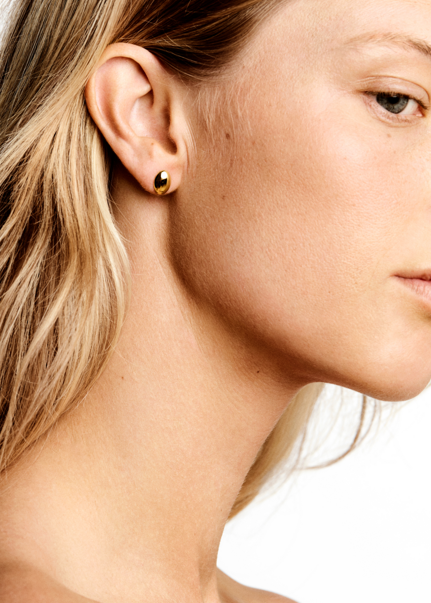 Close-up profile view of a person wearing a simple gold pair of Luna Earrings from Lie Studio on their left ear, with shoulder-length blonde hair and fair skin.