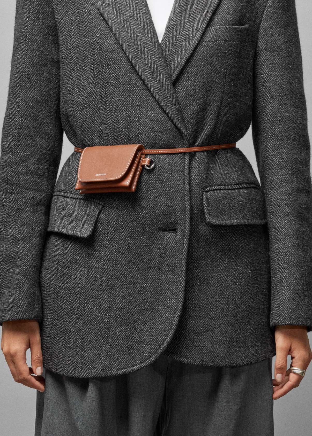 Someone wearing a grey herringbone blazer and trousers accessorized with the Lie Studio Stella Wallet, a small brown Italian cow leather belt bag, fastened around the waist for a versatile look.