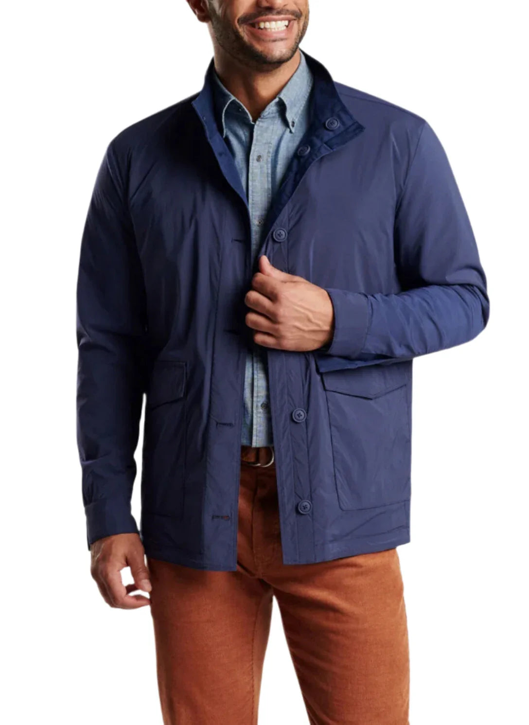 A man wearing a Peter Millar Stowe Reversible Safari Jacket in navy blue over a light blue shirt and brown pants, smiling as he adjusts the jacket with one hand.