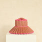 The Island Capri Hat by Lorna Murray—a multicolored woven sun hat featuring a wide, pleated brim and pink trim—sits on a white pedestal against a beige background. Perfect for travel, it’s expertly crafted to offer UPF 50+ protection.