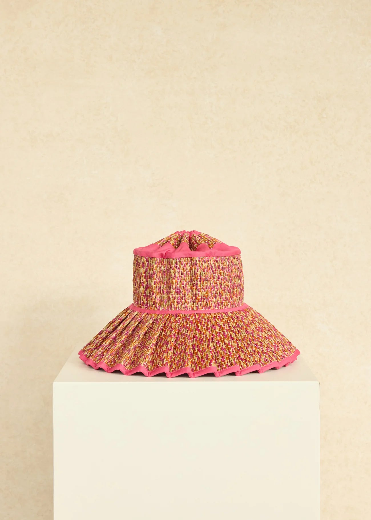 The Island Capri Hat by Lorna Murray—a multicolored woven sun hat featuring a wide, pleated brim and pink trim—sits on a white pedestal against a beige background. Perfect for travel, it’s expertly crafted to offer UPF 50+ protection.