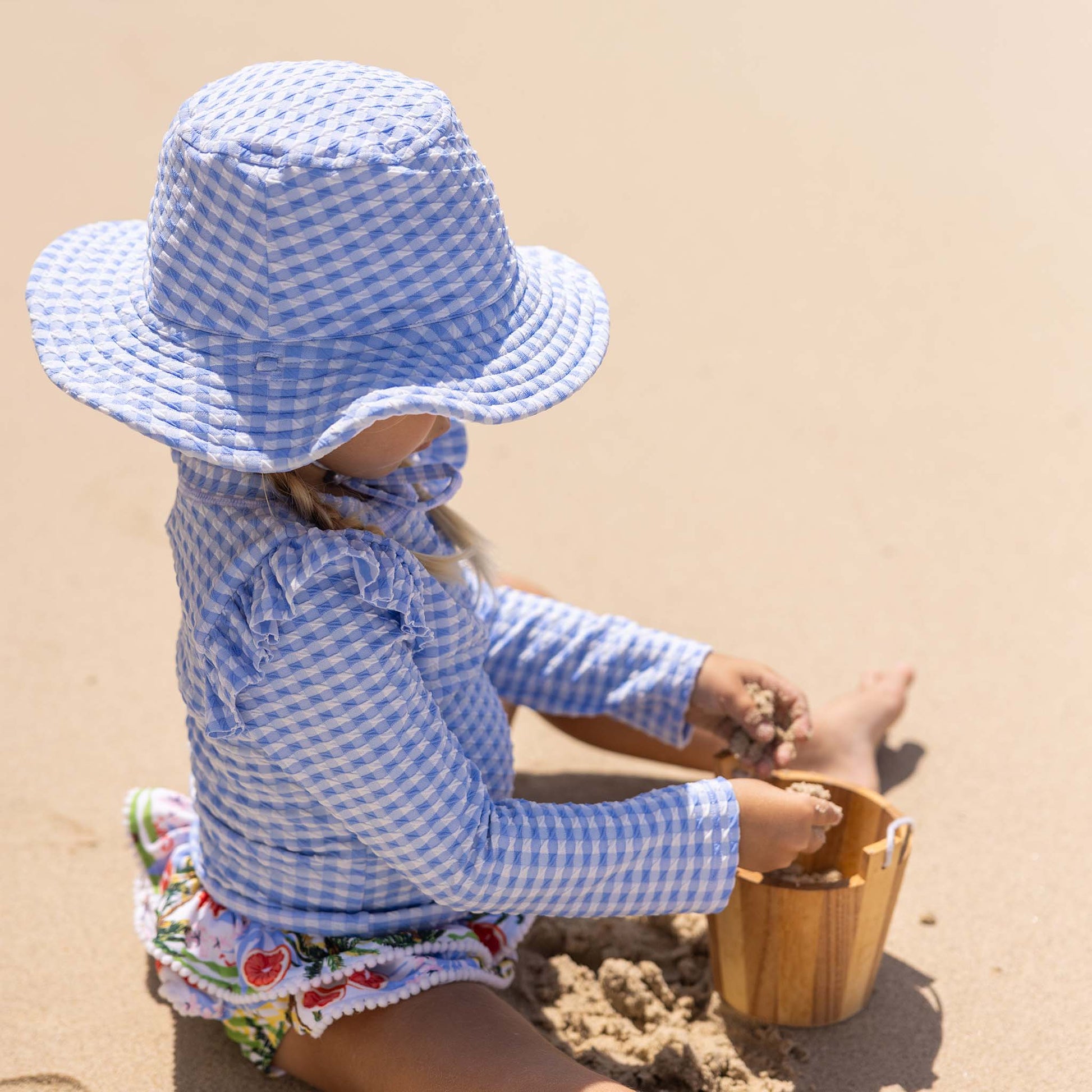 A young child wears the Snapper Rock Seaside Gingham Reversible Bucket Hat while sitting on the beach, dressed in a matching shirt and playing with sand using a small wooden bucket.