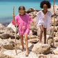 Two children wearing Snapper Rock Bubble Gum Beach Cover-Ups, featuring eyelet trim, walk with sticks on the rocky terrain near the ocean.