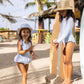 Two young girls wear Snapper Rock Baby Girls' Seaside Gingham Skirt Swimsuits and sun hats as they stand on a sandy beach near surfboards and a beach bar.