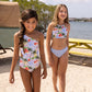Two girls wear Snapper Rock Girls' Ciao Paradiso One Shoulder Swimsuits on a sandy beach near a picnic table, with water and trees in the background.