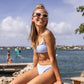 A girl wearing the Snapper Rock Girls' Seaside Gingham Teen Crop Bikini sits on a rock by the water, smiling in sunglasses, with windsurfers and others creating a lively seaside background.