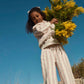 A young girl wears the Louise Misha Girls’ Ysalia Sweatpant, adorned with a delicate floral print, as she holds a large bouquet of yellow flowers outdoors beneath a clear blue sky.