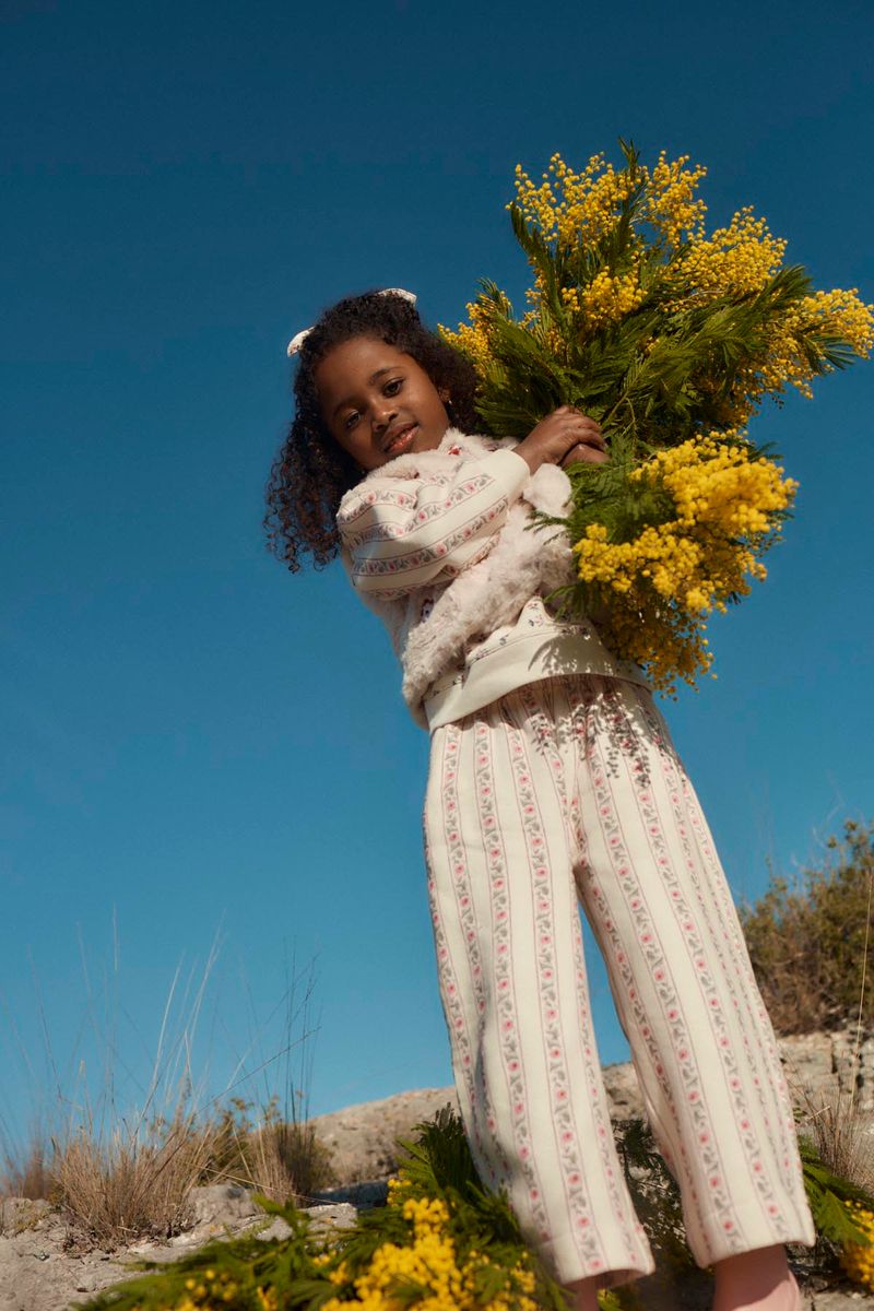 A young girl wears the Louise Misha Girls’ Ysalia Sweatpant, adorned with a delicate floral print, as she holds a large bouquet of yellow flowers outdoors beneath a clear blue sky.