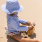 A young child wearing the Snapper Rock Baby Girls' Ciao Paradiso Ruffle Set made from recycled fabric plays with a wooden bucket on a sunny beach.