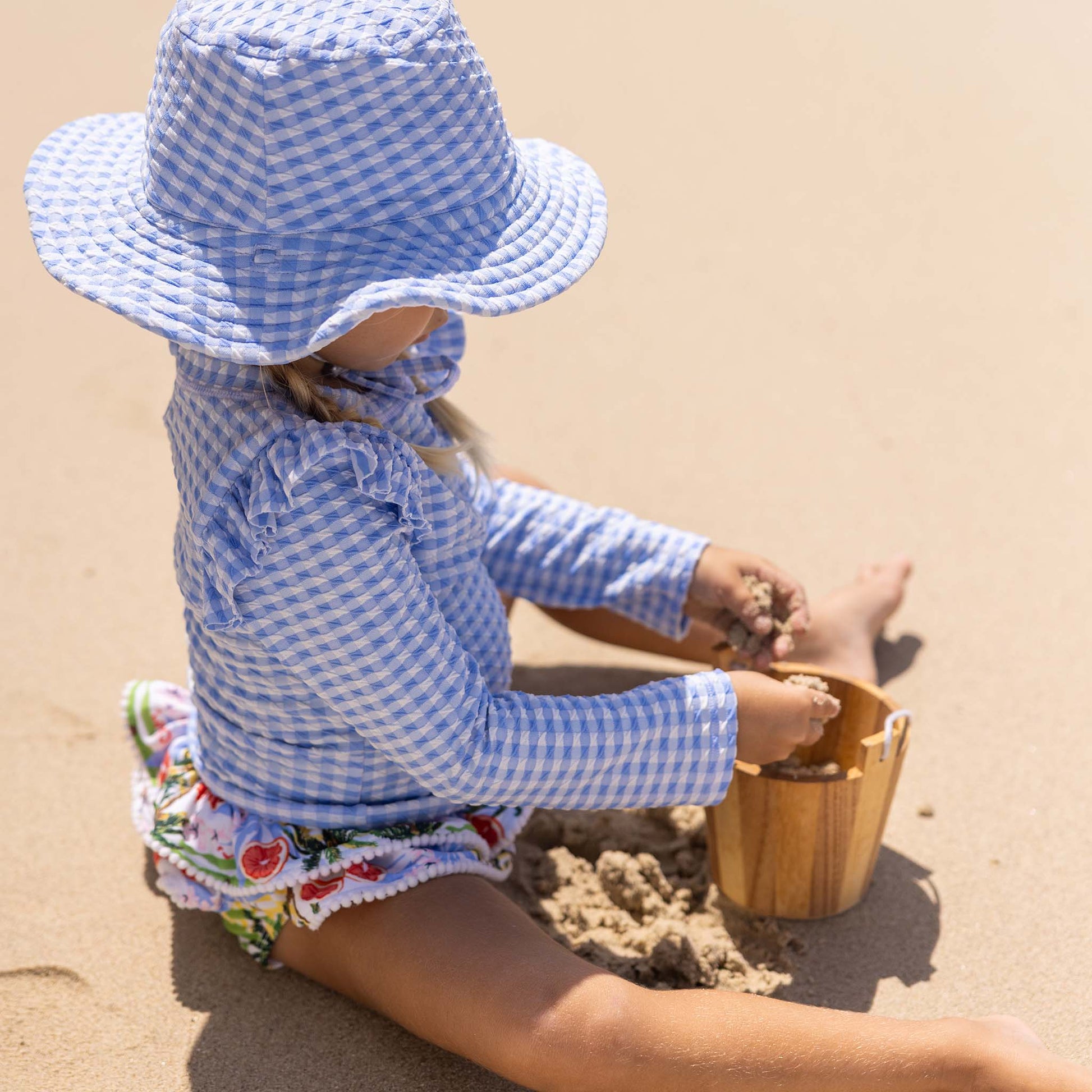 A young child wearing the Snapper Rock Baby Girls' Ciao Paradiso Ruffle Set made from recycled fabric plays with a wooden bucket on a sunny beach.