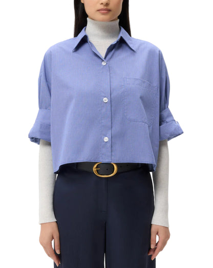 A woman stands facing forward, wearing the TWP Next Ex Stripe Shirt over a light turtleneck, paired with dark pants and a black belt with a gold buckle.