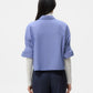 A woman faces away, wearing wide-leg navy pants, a light gray long-sleeve top, and the TWP Next Ex Stripe Shirt—a boxy, cropped blue button-down—layered over it.