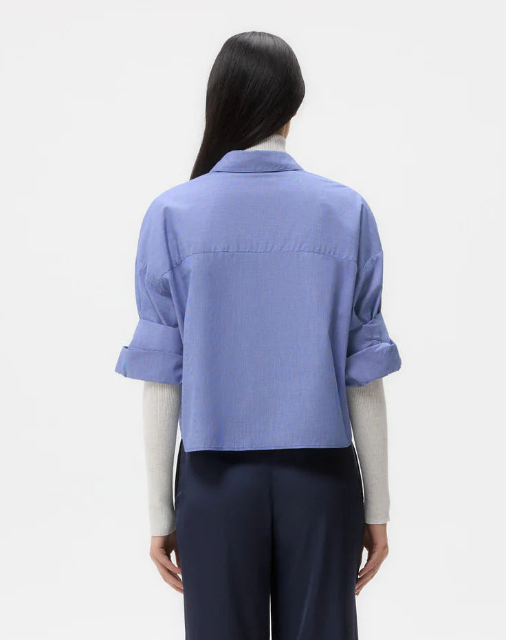 A woman faces away, wearing wide-leg navy pants, a light gray long-sleeve top, and the TWP Next Ex Stripe Shirt—a boxy, cropped blue button-down—layered over it.