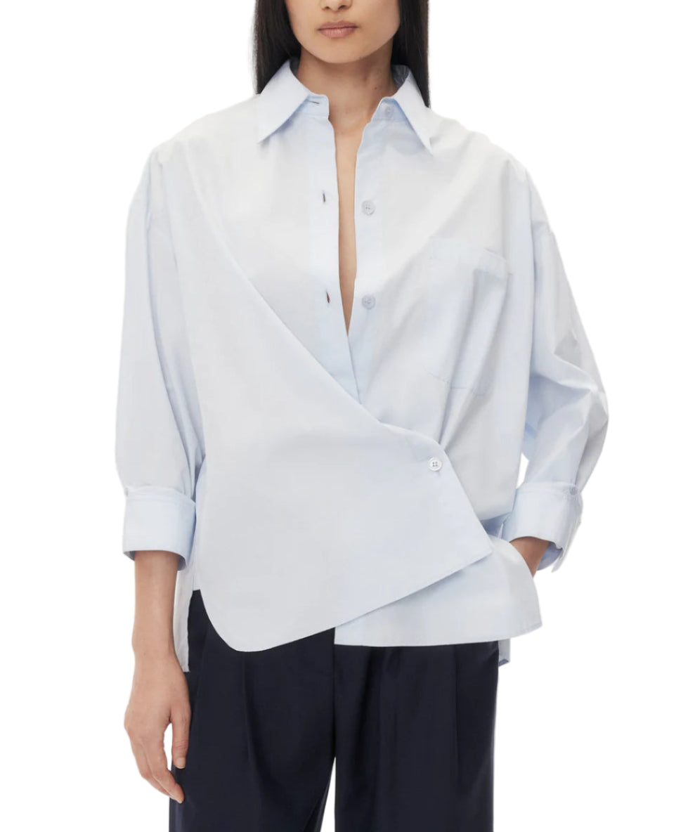 A person stands against a plain white background, looking directly at the camera, wearing the TWP New Earl Shirt—a relaxed, oversized, light blue button-down—paired with dark pants.