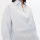 A woman wears the TWP New Earl Shirt, a relaxed, light blue oversized button-down with an asymmetrical design, standing against a plain white background.