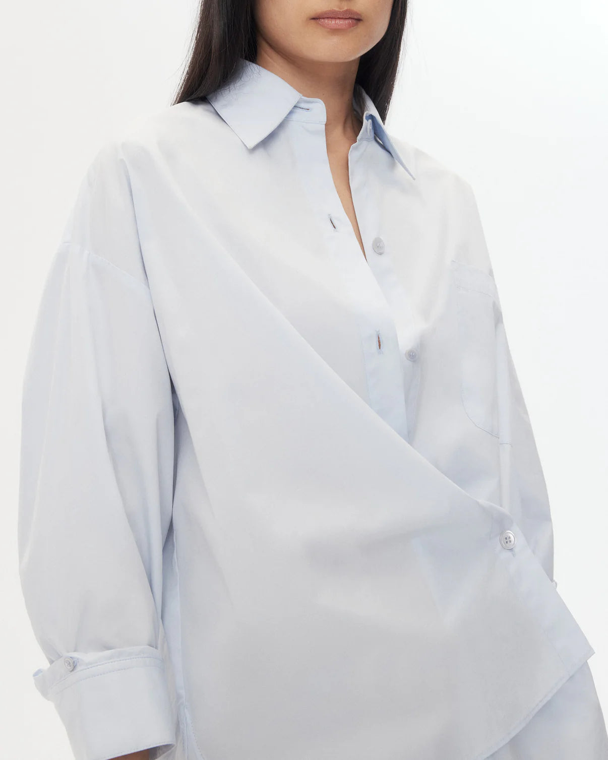 A woman wears the TWP New Earl Shirt, a relaxed, light blue oversized button-down with an asymmetrical design, standing against a plain white background.