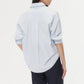 A person stands with their back to the camera wearing the TWP New Earl Shirt in light blue, featuring rolled sleeves, paired with dark pants against a plain white background.
