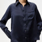 A woman stands against a plain background wearing the TWP Joey Silk Shirt, a navy blue long-sleeve classic fit top with a chest pocket.