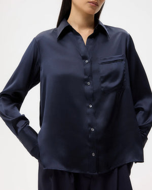 A woman stands against a plain background wearing the TWP Joey Silk Shirt, a navy blue long-sleeve classic fit top with a chest pocket.