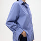 A woman poses against a plain background in the TWP Next Morning Top, a relaxed silhouette shirt in light blue cotton poplin, paired with dark jeans and one hand in her pocket.