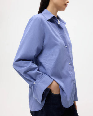 A woman poses against a plain background in the TWP Next Morning Top, a relaxed silhouette shirt in light blue cotton poplin, paired with dark jeans and one hand in her pocket.