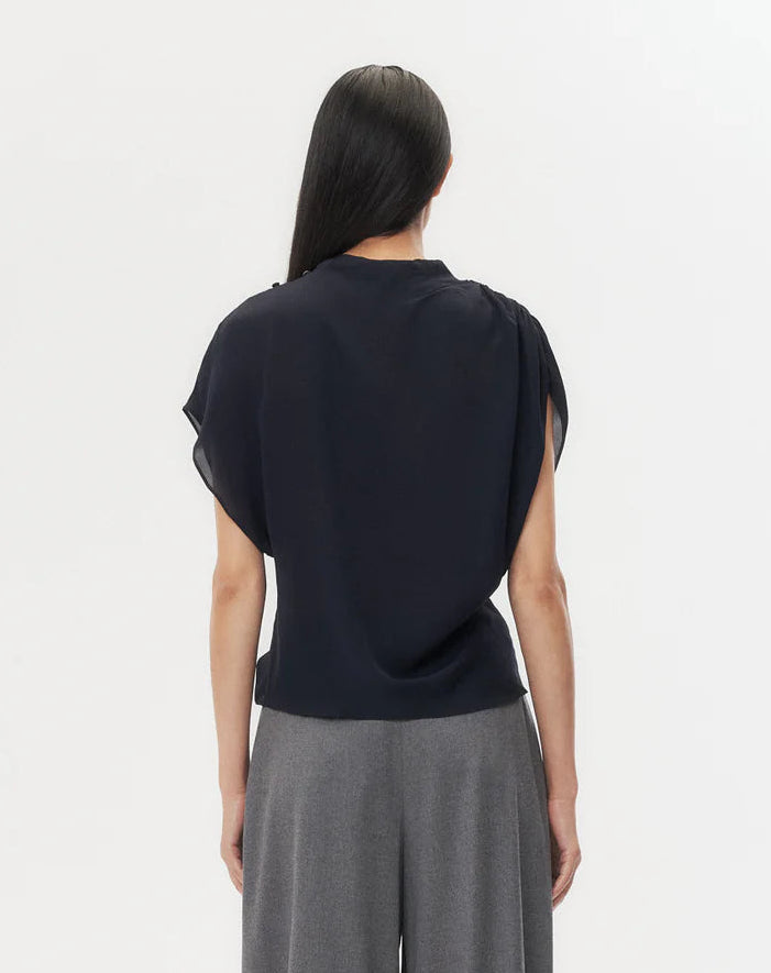 A person with long dark hair stands facing away, wearing the TWP Wish You Would Top and gray wide-leg pants against a plain white background.