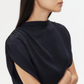 A woman with long dark hair wears the loose-fitting Wish You Would Top by TWP, featuring cap sleeves, and stands with arms crossed against a plain light background.