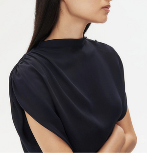 A woman with long dark hair wears the loose-fitting Wish You Would Top by TWP, featuring cap sleeves, and stands with arms crossed against a plain light background.