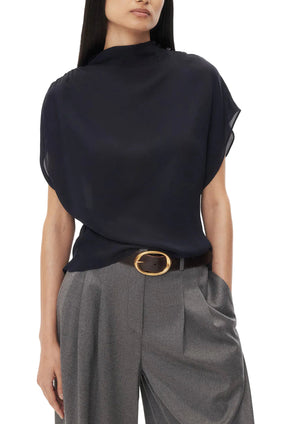 A person wearing the TWP Wish You Would Top, a loose-fitting navy mock neck blouse with short sleeves, paired with high-waisted gray pleated trousers and a wide belt with a large gold buckle.