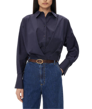 A woman wearing the TWP Taken Shirt, a relaxed navy button-down, tucked into high-waisted blue jeans with a brown belt, stands against a white background.