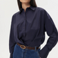A person wearing the TWP Taken Shirt—a relaxed navy cotton poplin button-down—tucked into high-waisted blue jeans with a brown belt.