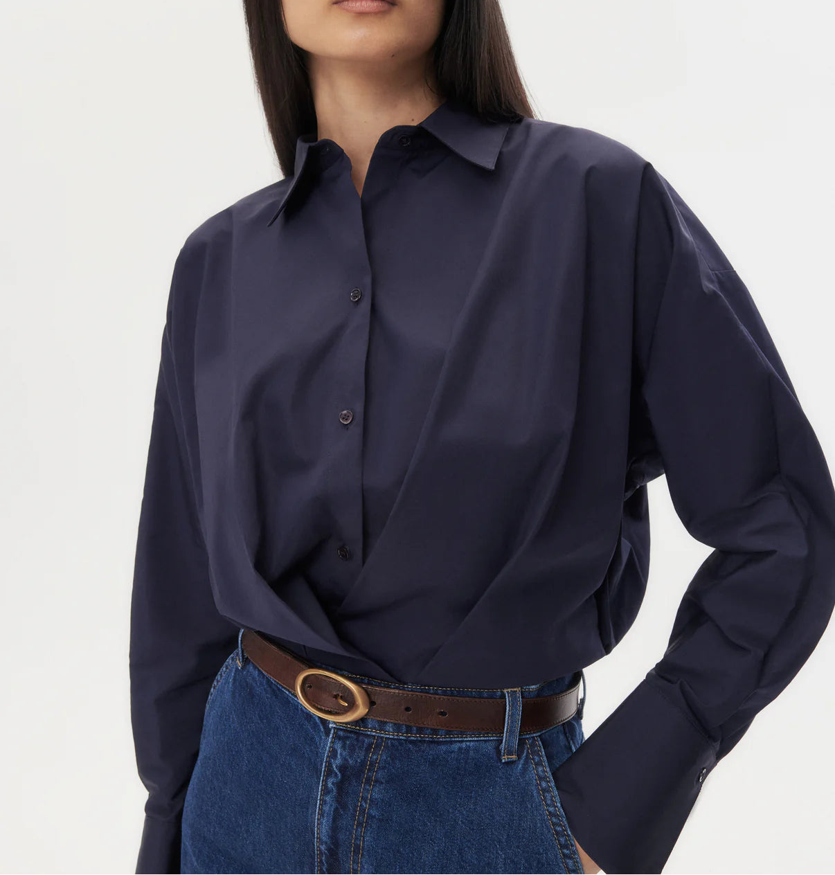 A person wearing the TWP Taken Shirt—a relaxed navy cotton poplin button-down—tucked into high-waisted blue jeans with a brown belt.