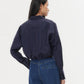 A woman with long dark hair models the TWP Taken Shirt, a relaxed navy cotton poplin button-down, paired with blue jeans, standing with her back to the camera against a plain white background.