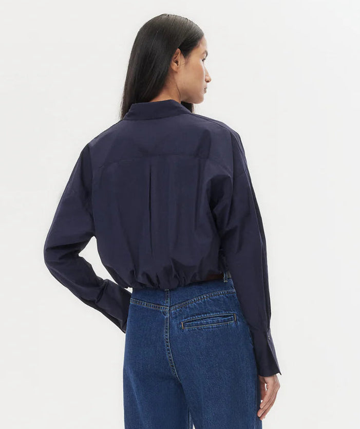 A woman with long dark hair models the TWP Taken Shirt, a relaxed navy cotton poplin button-down, paired with blue jeans, standing with her back to the camera against a plain white background.