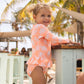 A young child wears the Snapper Rock Baby Girls' Cali Palms Recycled Skirt Surf Suit, standing by a light wooden table outdoors with palm trees and shade structures in the background.